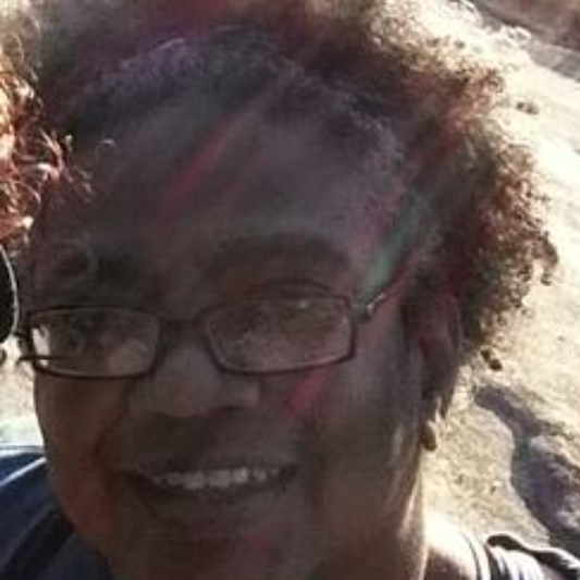 Photo of Jackie Marshall, an African American woman wearing glasses, a neat hairstyle and smiling with sun rays combing through her hair