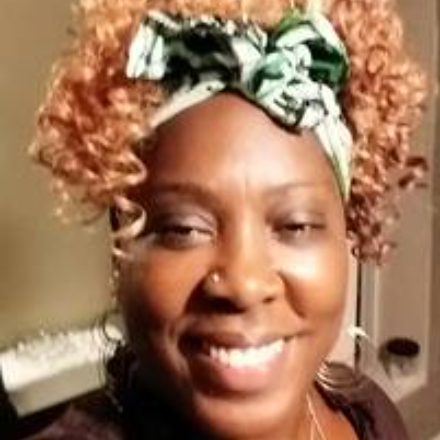 Photo of Doctor Hadrakel Reuven, an African American woman with light brown, almost red colored curled hair, wearing a headwrap tied in a bow, smiling